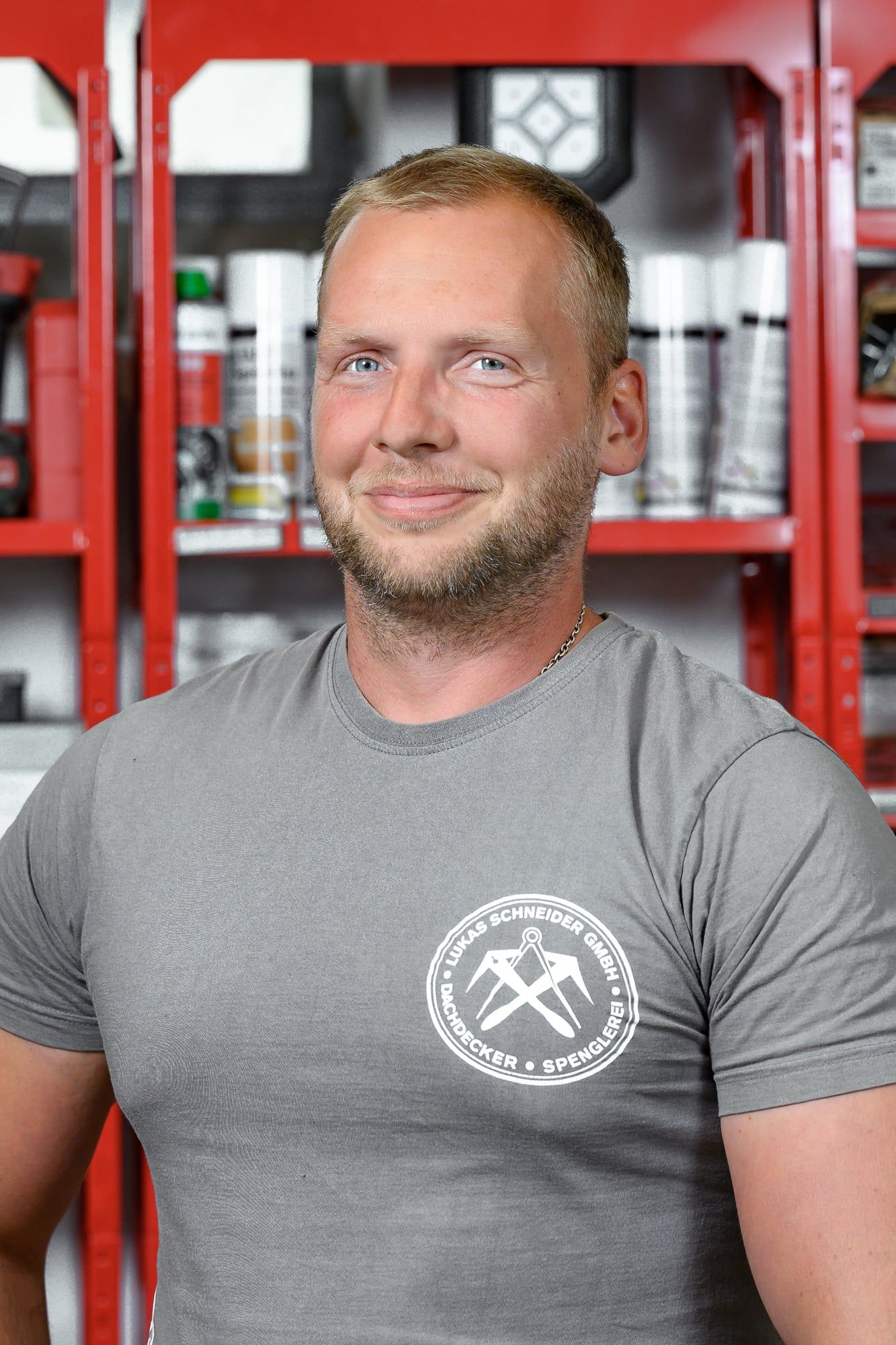 Portrait eines Dachdeckers der Dachdeckerei Lukas Schneider im grauen Firmenshirt vor Werkstattregalen – Businessfotos in München