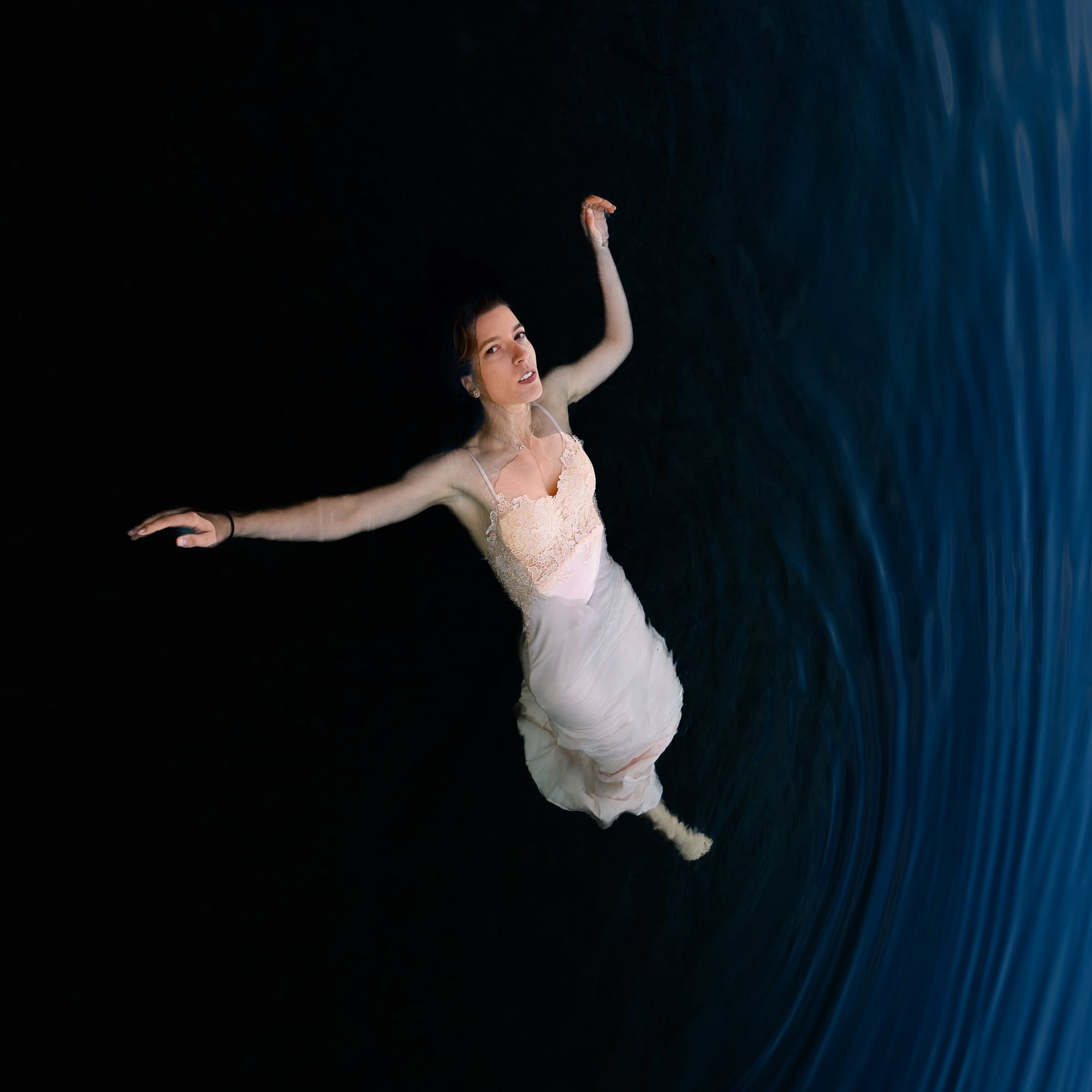 Frau im hellen Kleid schwebt mit ausgebreiteten Armen auf dunkelblauem Wasser, von oben fotografiert – Portrait in Augsburg
