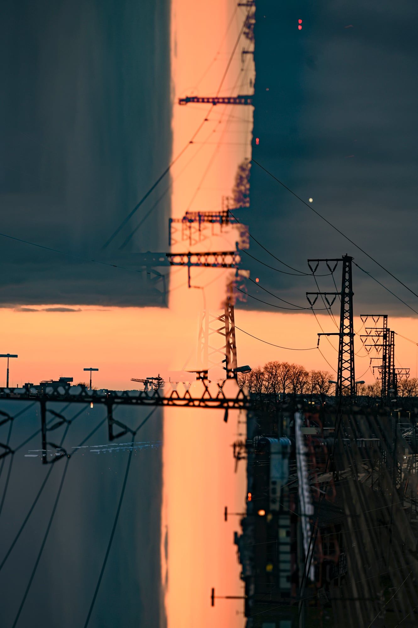 Mehrfachbelichtung im Hochformat: Gleise und Strommasten kreuzen sich vor orangem Himmel – Augsburg
