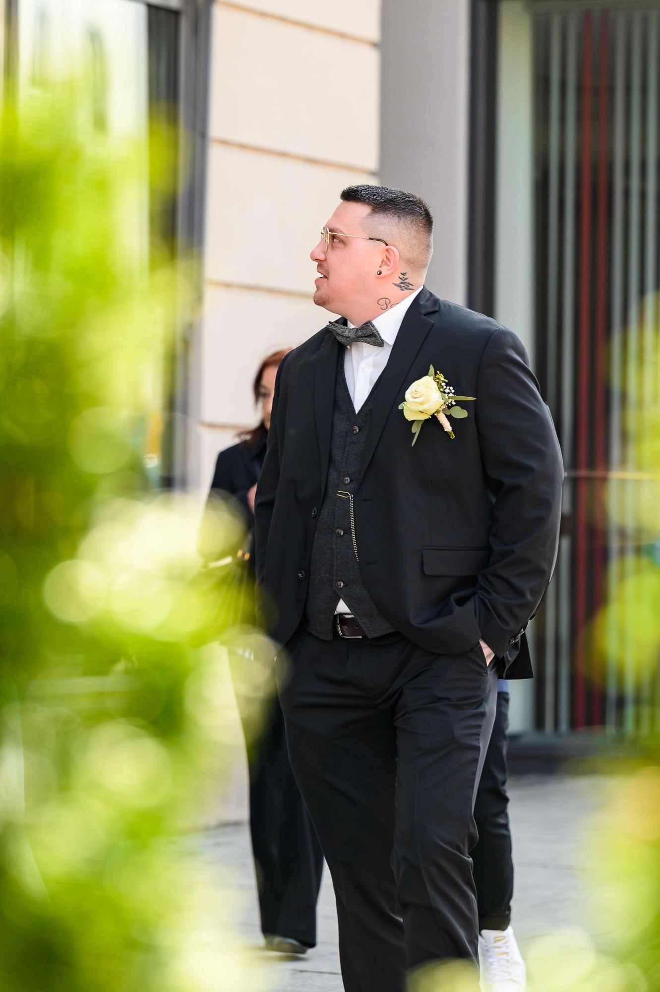 Bräutigam im dunklen Anzug wartet vor einem Gebäudeeingang, im Vordergrund unscharfes Grün – Hochzeit in Augsburg