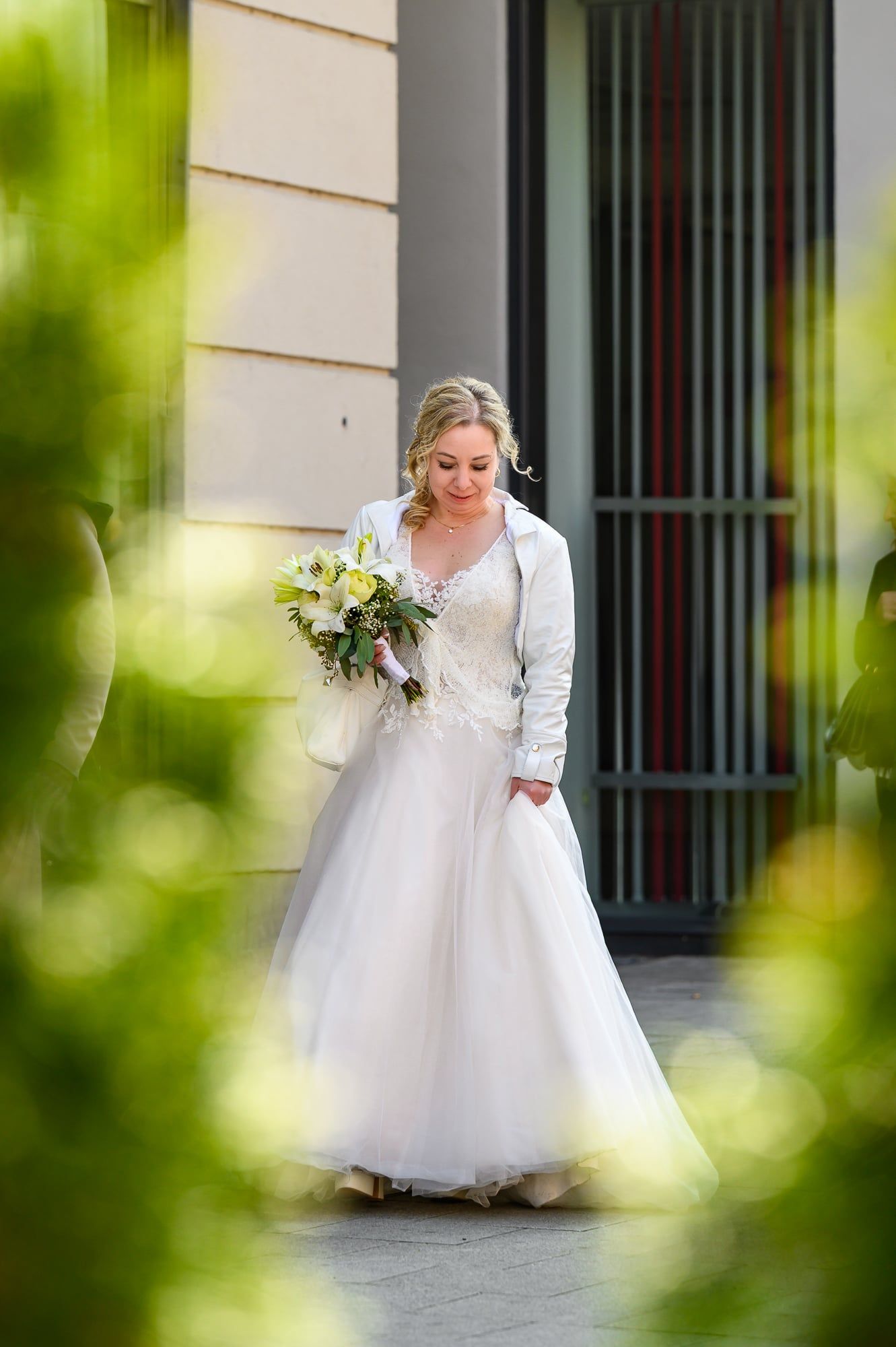 Braut im weißen Kleid mit Brautstrauß tritt aus einem Eingang, durch Grün hindurch fotografiert – Hochzeit in Augsburg
