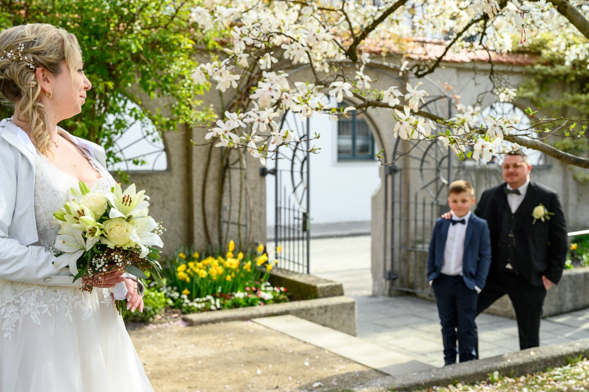 Braut mit Strauß blickt zu Bräutigam und Kind, die durch einen blühenden Garten kommen – Hochzeit in Augsburg
