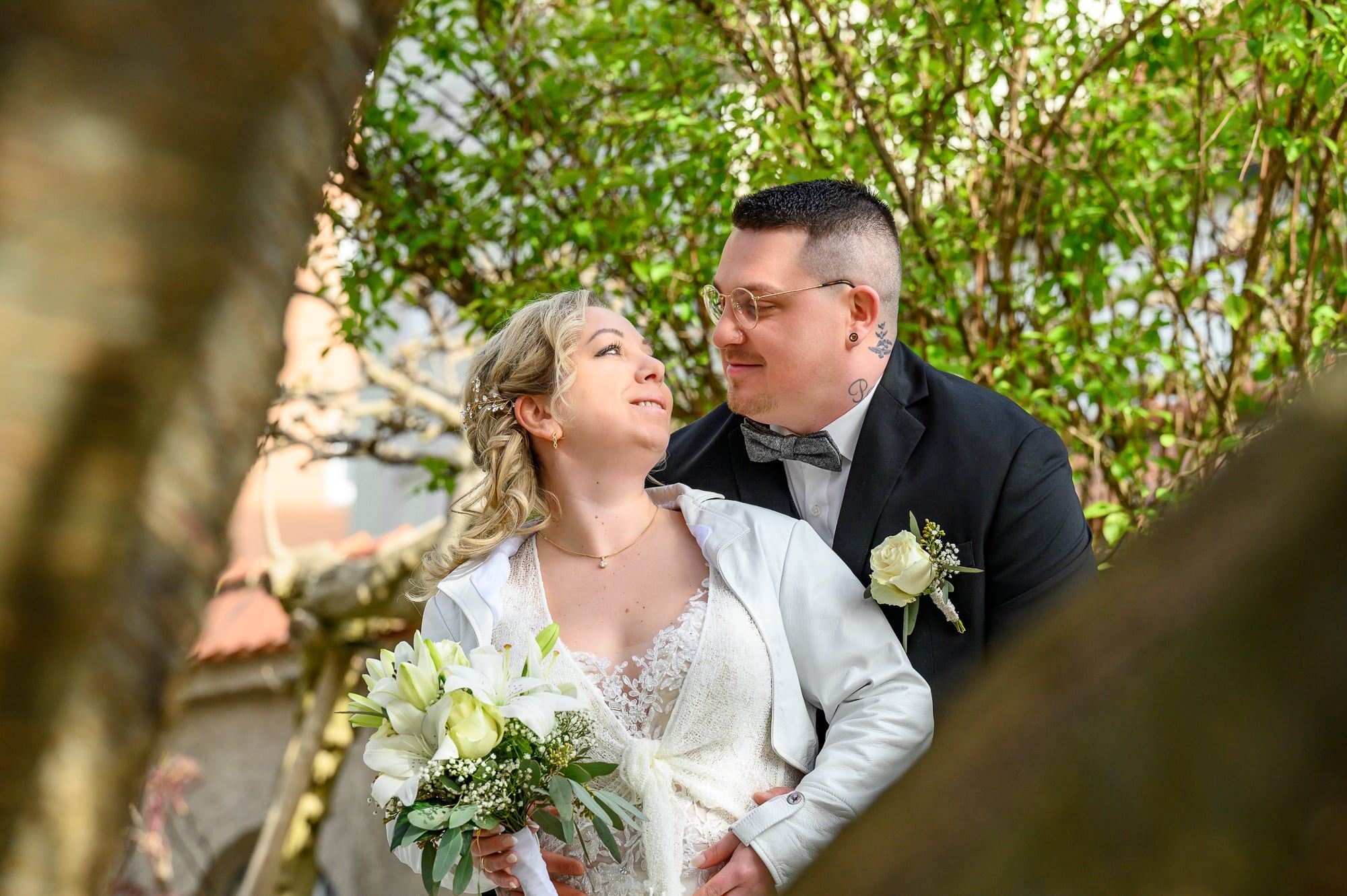 Brautpaar lacht sich in einem blühenden Garten an, durch Zweige hindurch fotografiert – Hochzeit in Augsburg