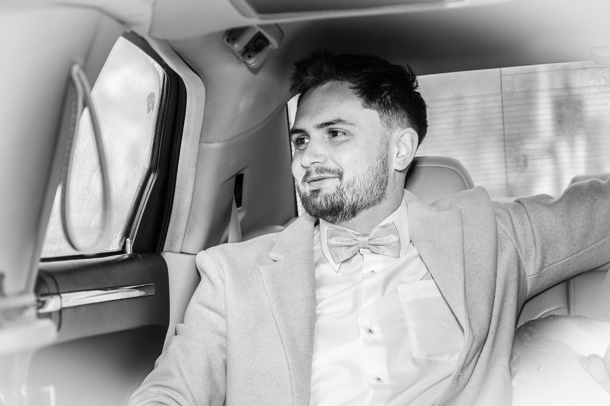 Bräutigam mit Fliege lehnt lächelnd im Hochzeitsauto, Schwarzweiß – Hochzeit in Augsburg