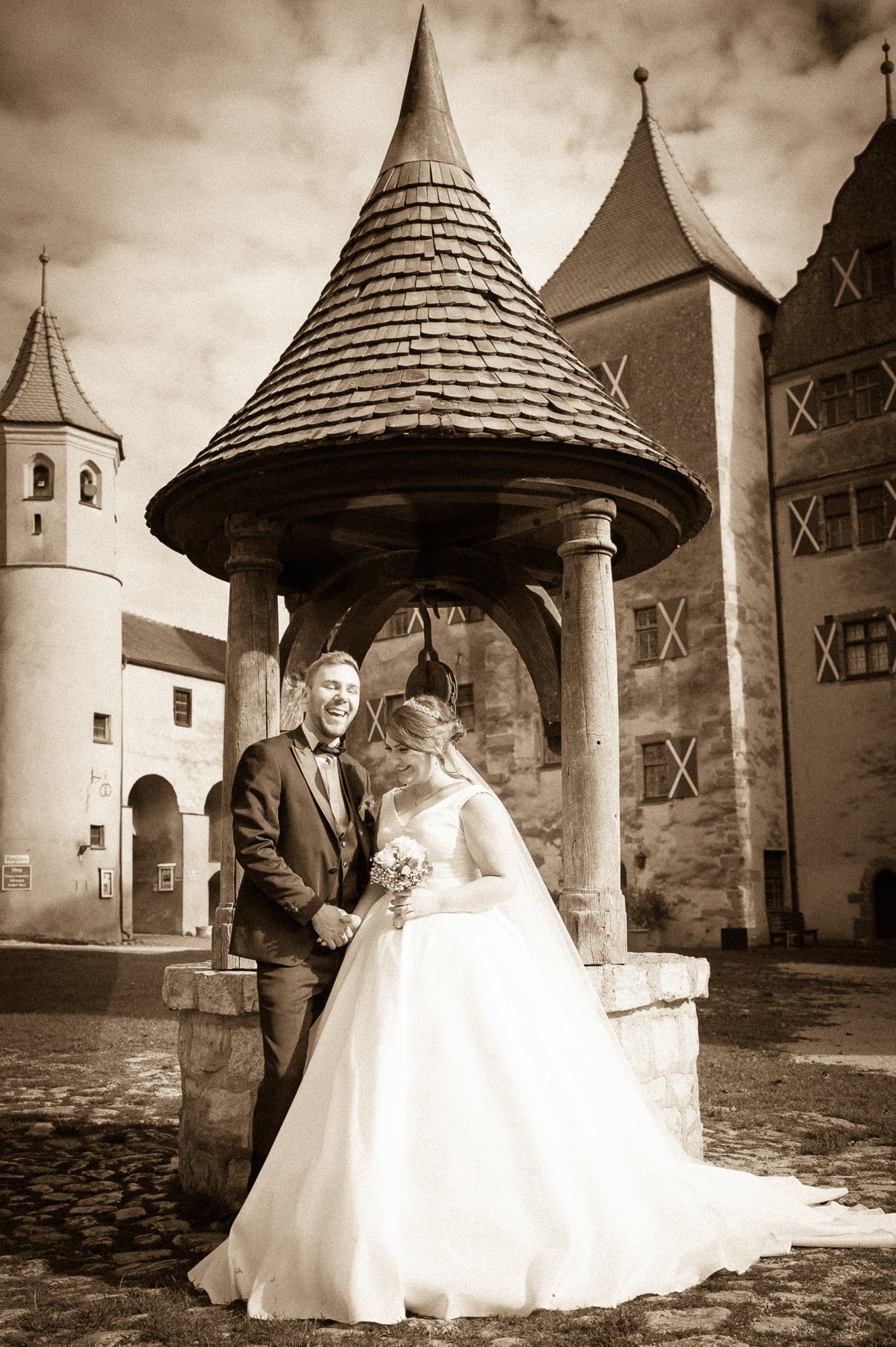 Brautpaar vor einem Brunnenhäuschen mit spitzem Dach in einem Schlosshof, in Sepia – Hochzeit in Wittislingen