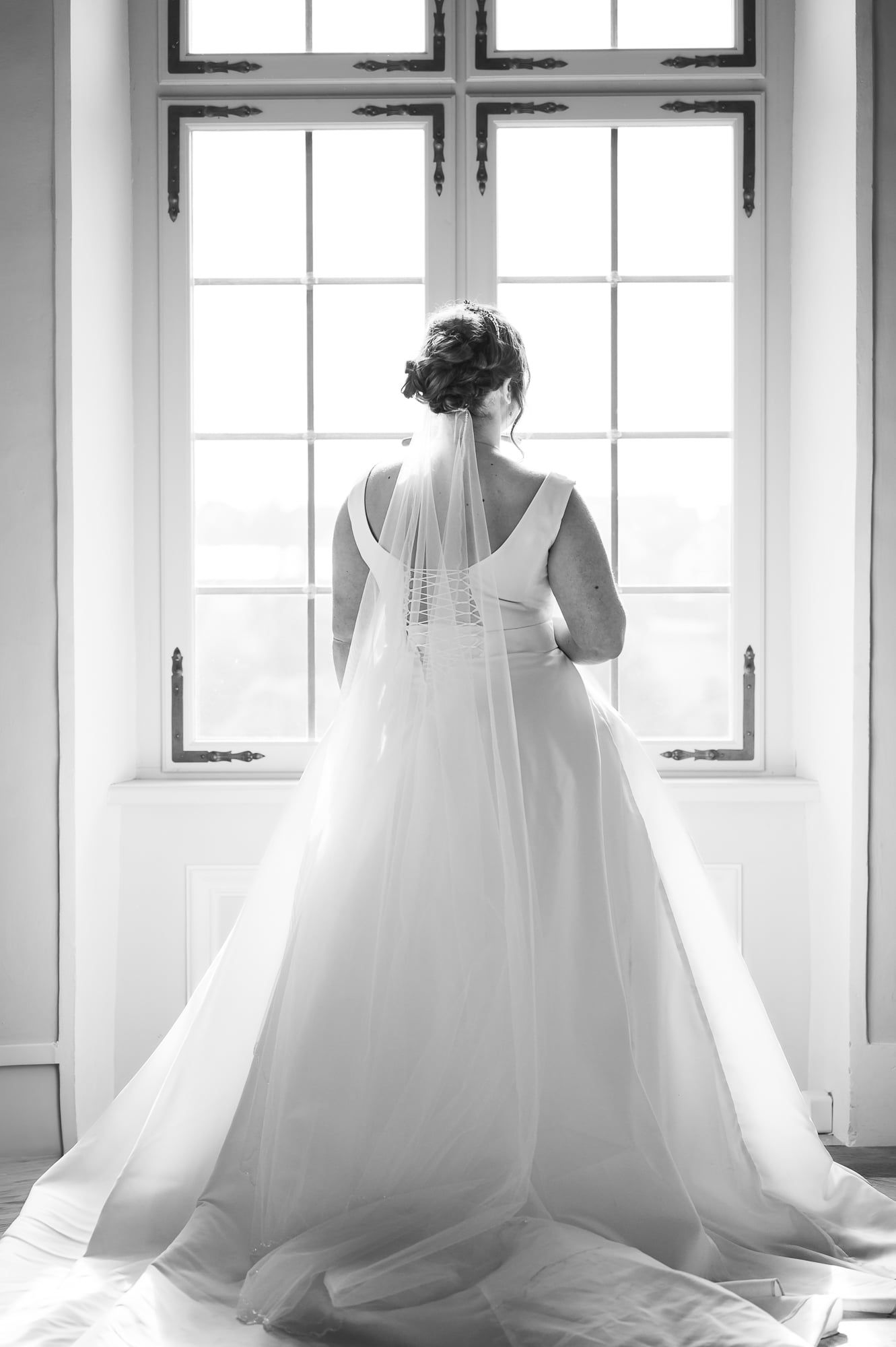 Braut im langen Kleid mit Schleier steht mit dem Rücken zur Kamera vor einem hohen Fenster – Hochzeit in Wittislingen