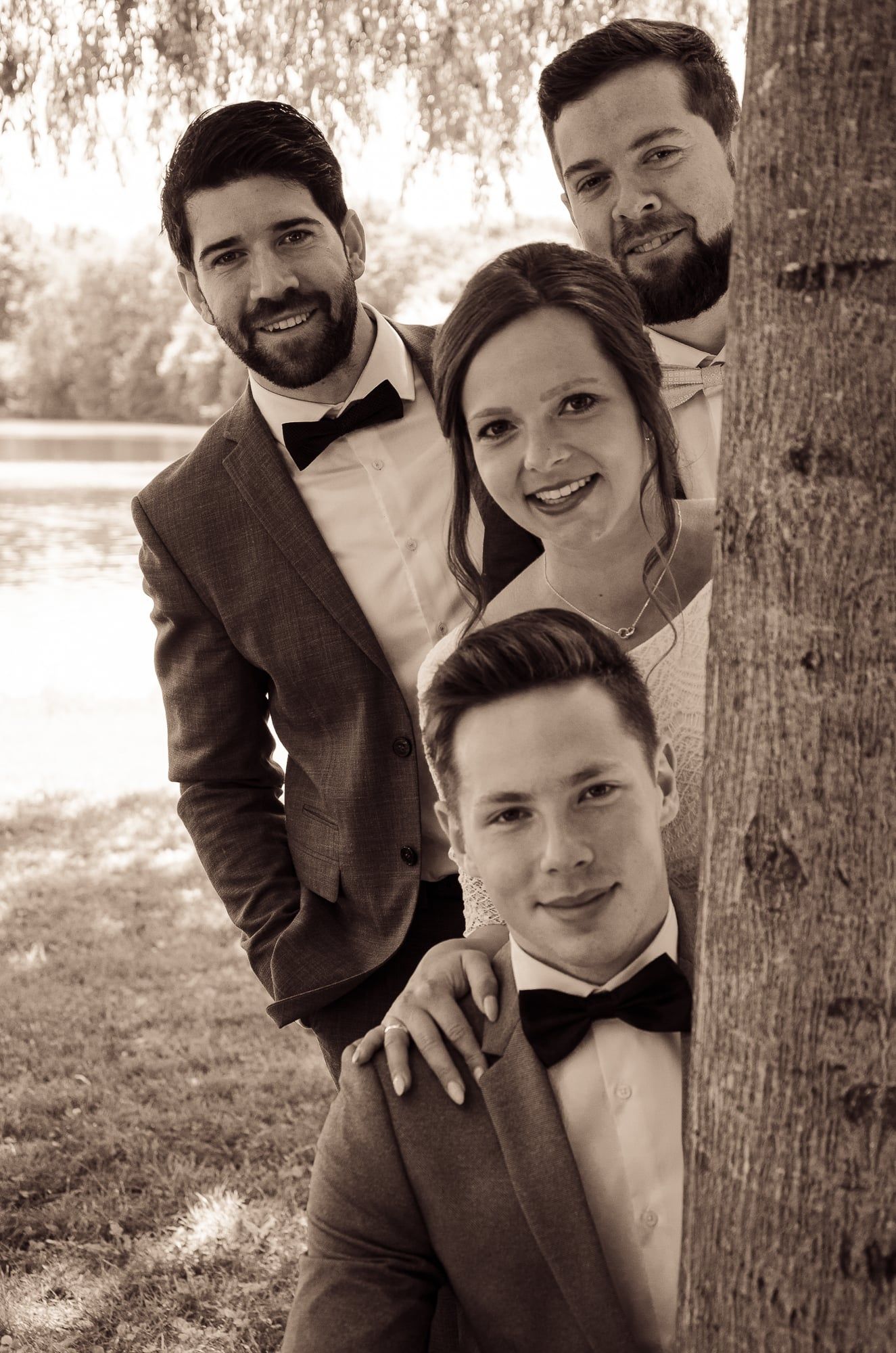 Brautpaar und zwei Freunde schauen hinter einem Baumstamm hervor, in Sepia – Hochzeit in Augsburg