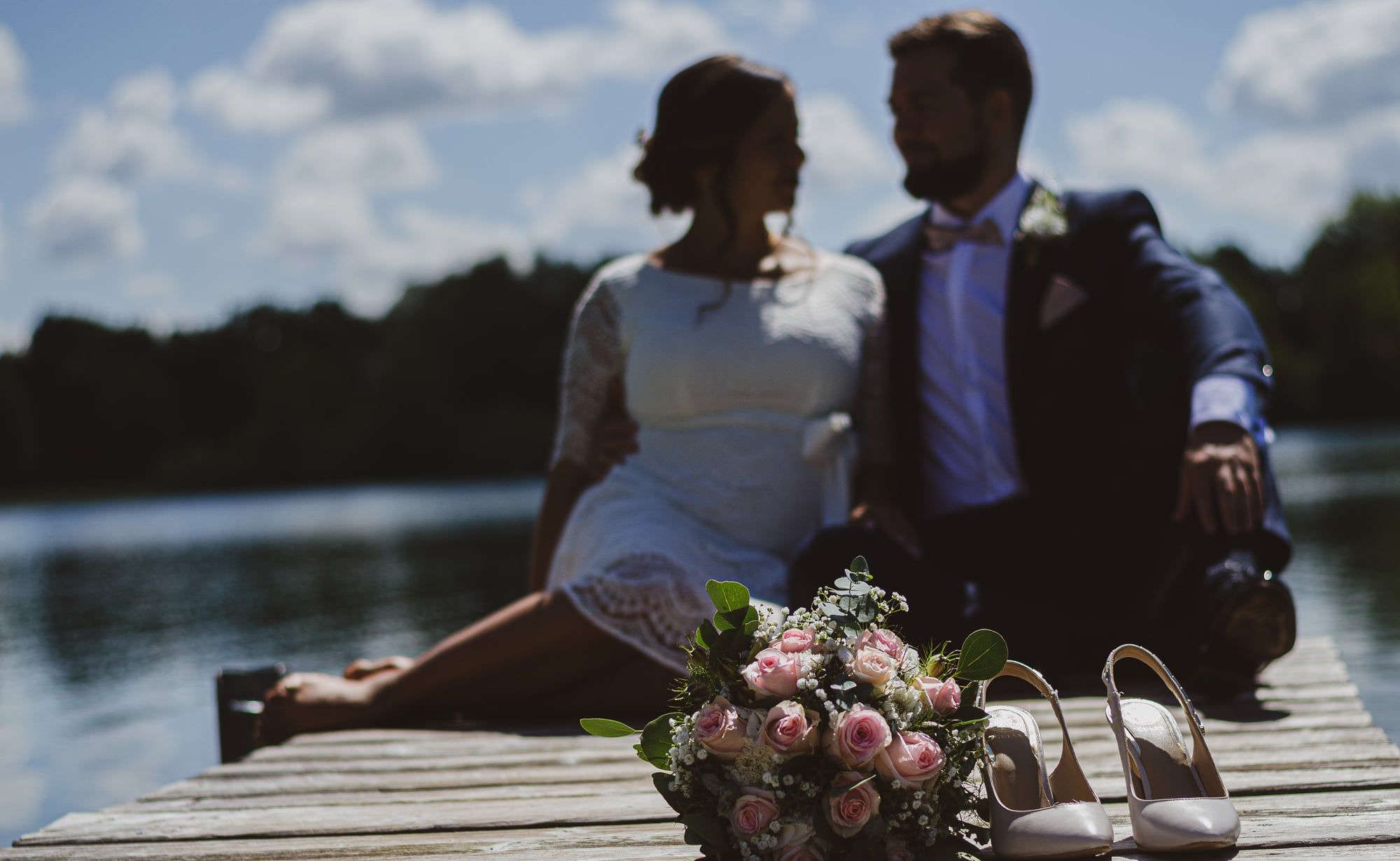Brautpaar sitzt auf einem Steg am See, davor Brautstrauß und Schuhe – Hochzeit in Augsburg