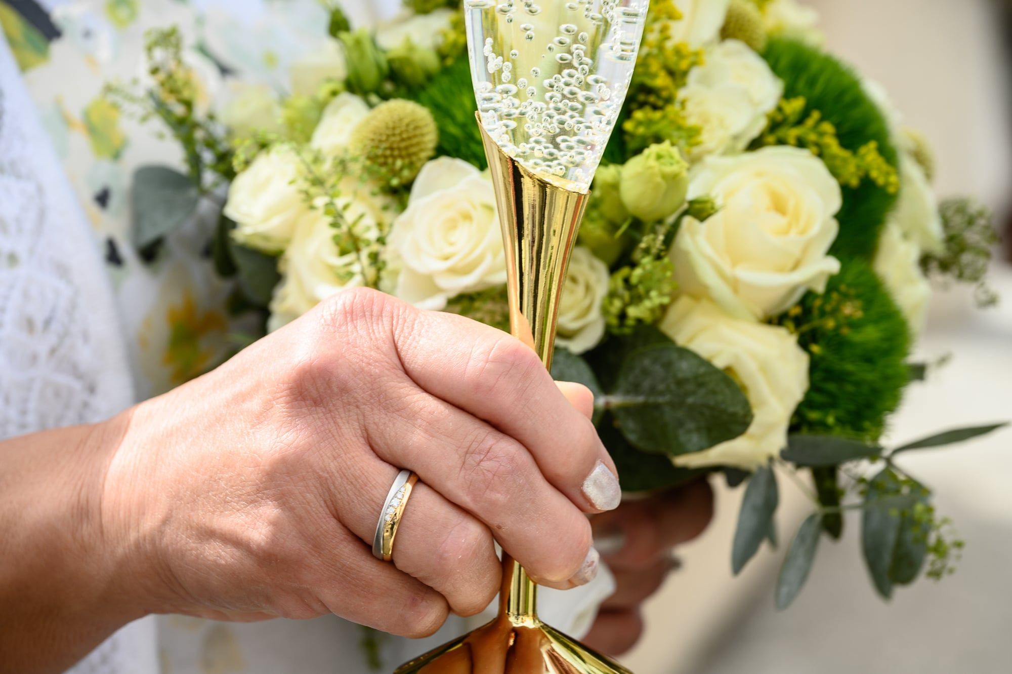 Hand mit Ehering hält ein Sektglas vor einem Brautstrauß aus weißen Rosen – Hochzeit in Friedberg