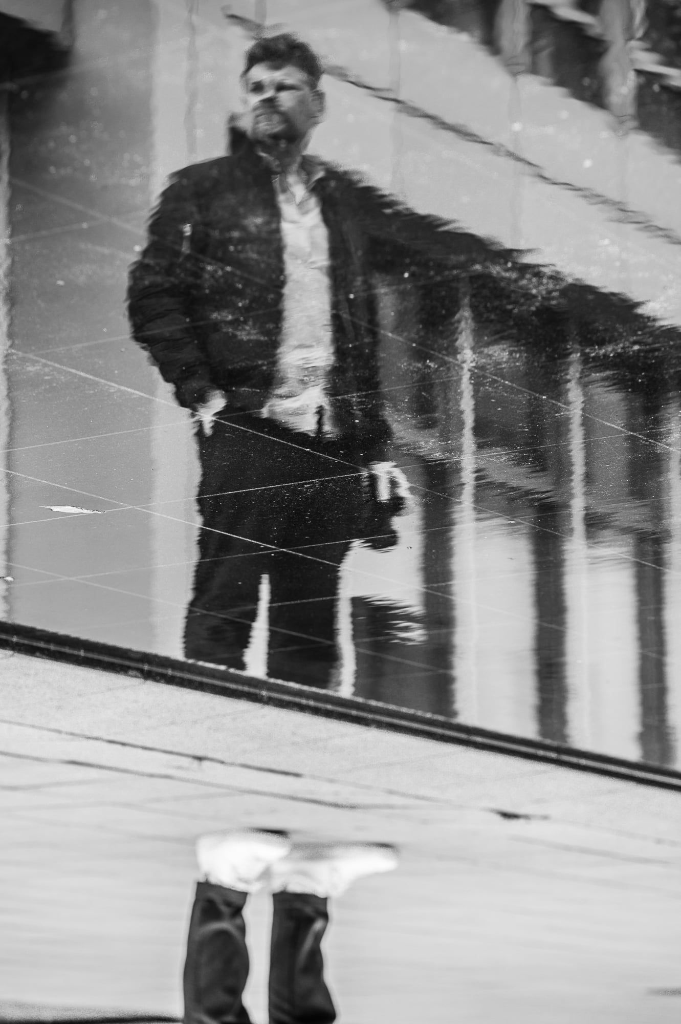 Mann steht auf einer spiegelnden Fläche, in der sich eine Hausfassade spiegelt, Schwarzweiß – Portrait in München