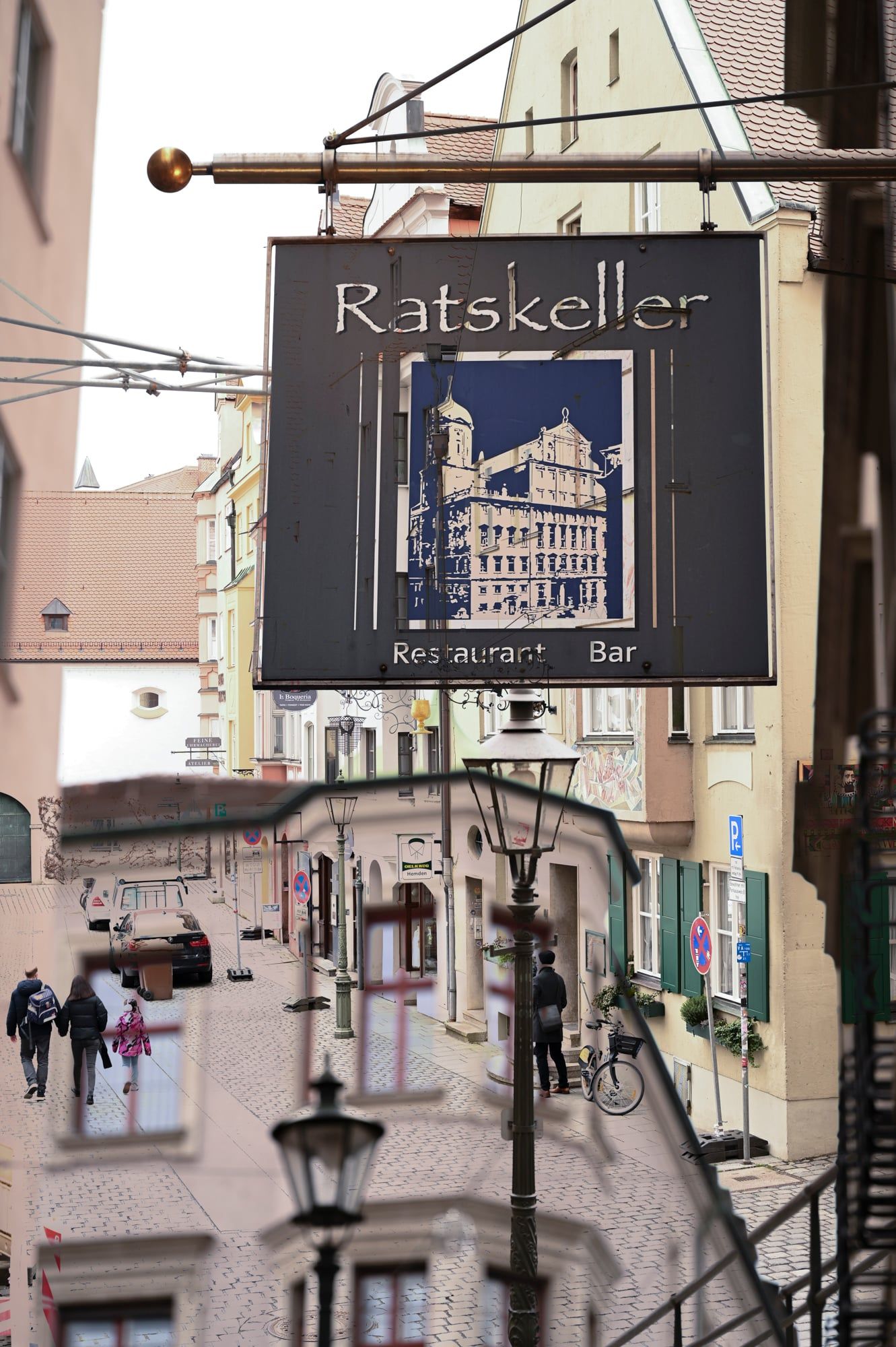 Schild des Ratskellers mit Rathaus-Motiv, darunter Passanten in der Gasse – Augsburg