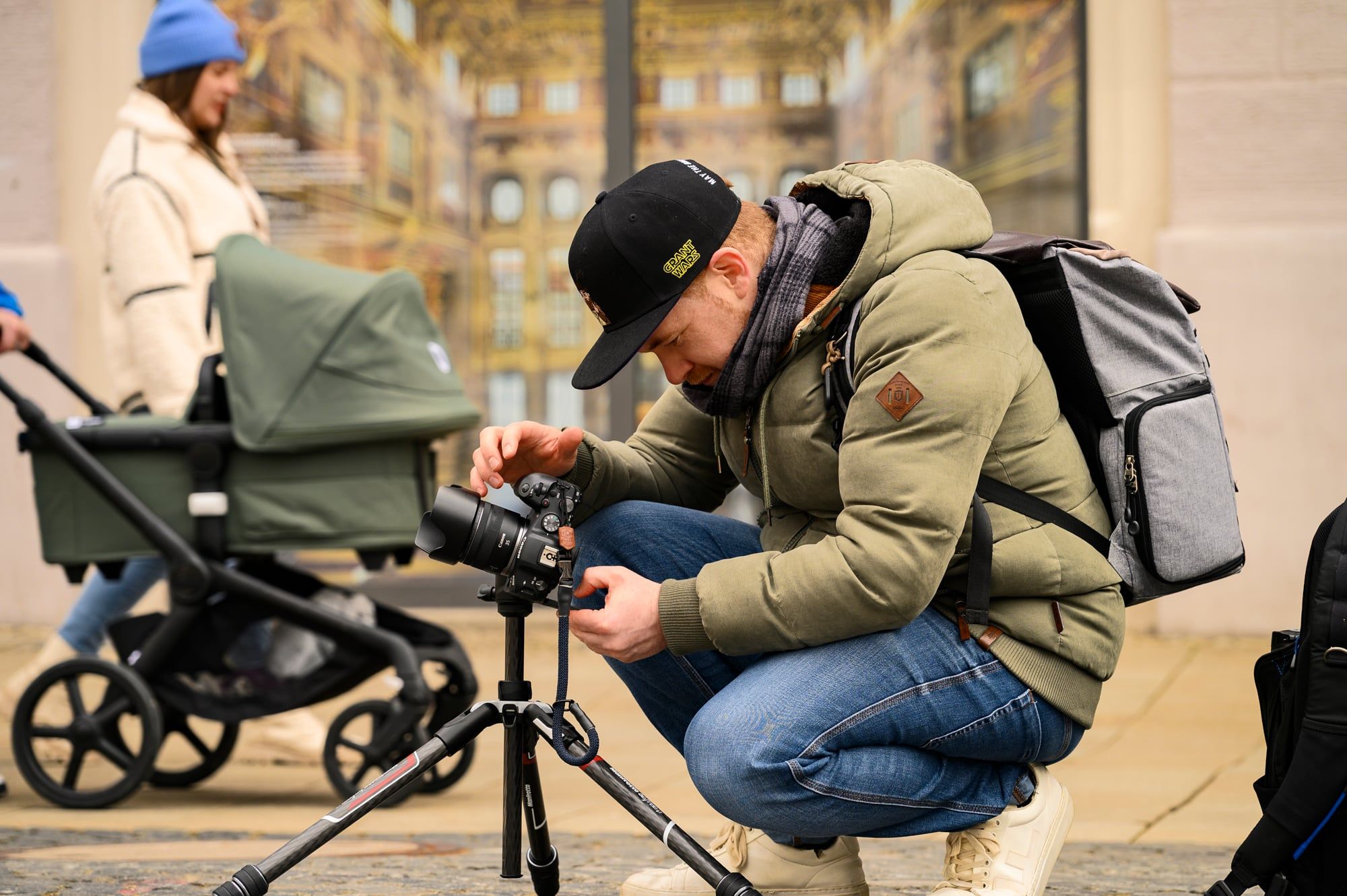 Daniel Köberle kniet mit Kamera und Stativ auf dem Augsburger Rathausplatz, hinter ihm eine Frau mit Kinderwagen