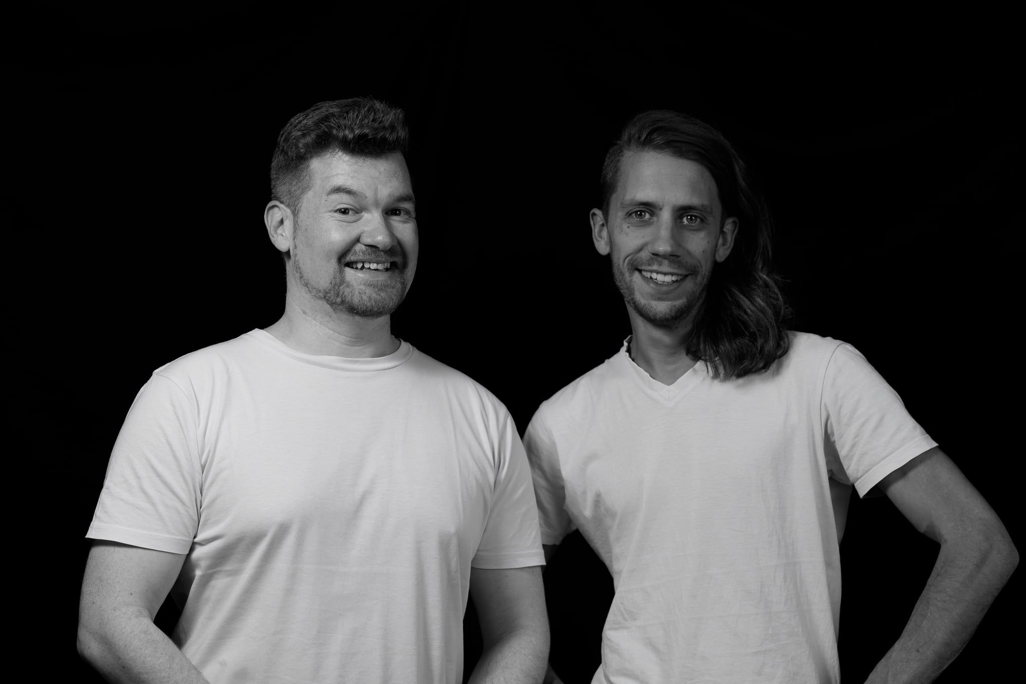 Daniel und Lukas lachend nebeneinander im Low-Key-Licht vor schwarzem Hintergrund